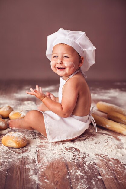 Menina encantadora com chapéu de cozinheiro e avental sentado na farinha com pães rindo alegremente
