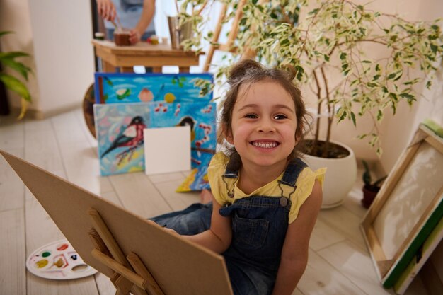 Menina encantadora de camiseta amarela e jeans azul, segurando um pincel e pintando em tela usando tintas aquarela ou óleo, sentada no chão ao lado de fotos pintadas em casa Conceito de arte infantil