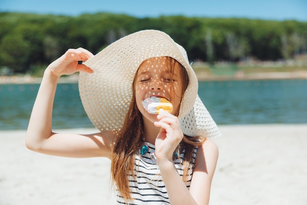 Menina encantadora de chapéu come sorvete na praia da praia Conceito de férias de verão