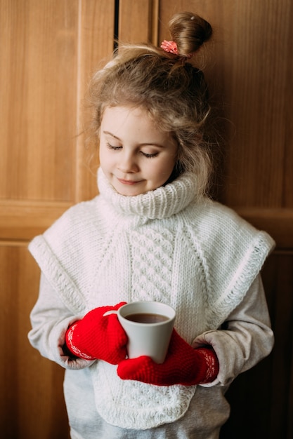 Menina encantadora de sete anos bebe chá quente, em pé ao lado da janela. ela está vestindo um suéter de tricô quente, suas mãos estão em luvas de tricô vermelhas. época de natal.