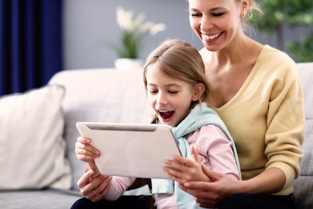 Menina encantadora e sua linda jovem mãe usando tablet digital e sorrindo enquanto está sentada no sofá em casa