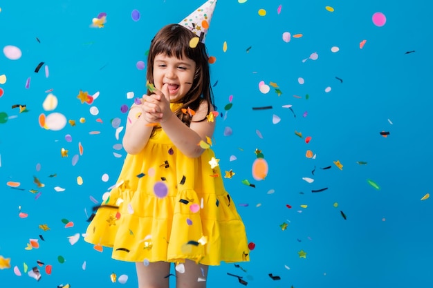 Menina encantadora em um vestido amarelo sobre fundo azul lança confete colorido aniversário