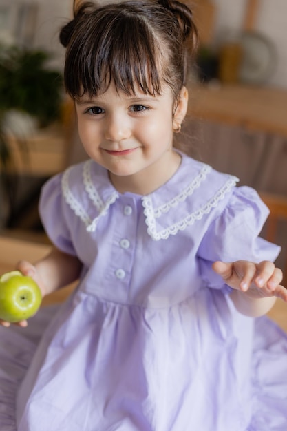 menina encantadora em um vestido lilás come uma maçã verde na cozinha. espaço para texto, banner