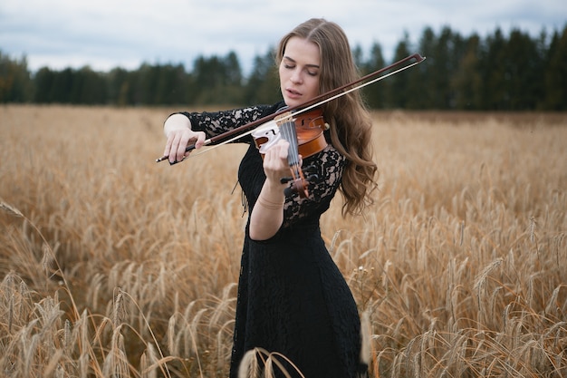 Menina encantadora em um vestido preto tocando violino com entusiasmo em um campo de trigo