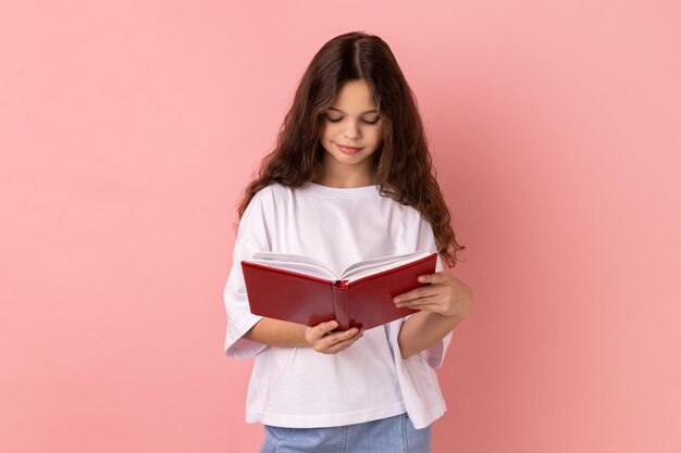 Menina encantadora encantada segurando nas mãos e lendo um livro interessante sendo muito atenciosa