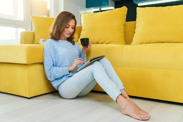 Menina encantadora segura seu computador tablet nas mãos sentada no chão perto de sofá amarelo moderno e