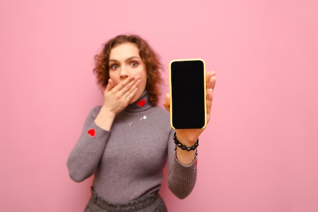 Menina encaracolada chocada cobriu a boca com a mão, segurando um smartphone com uma tela preta na mão