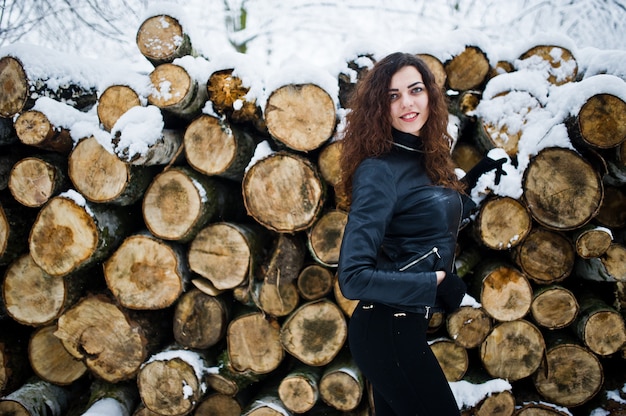 Menina encaracolada de elegância no casaco de cabedal no parque florestal nevado no inverno contra tocos.