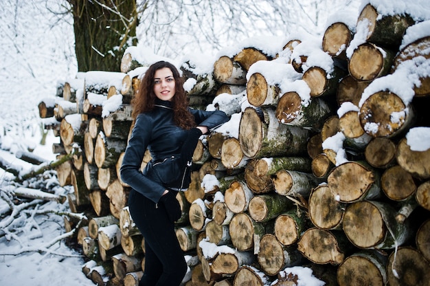 Menina encaracolada de elegância no casaco de cabedal no parque florestal nevado no inverno contra tocos.