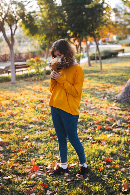 Menina encaracolada de suéter amarelo na grama com buquê de outono de folhas secas e flores