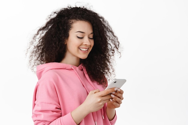 Foto menina encaracolada moderna mandando mensagens de texto, conversando no smartphone, sorrindo e olhando para o celular, usando o aplicativo, em pé contra um fundo branco