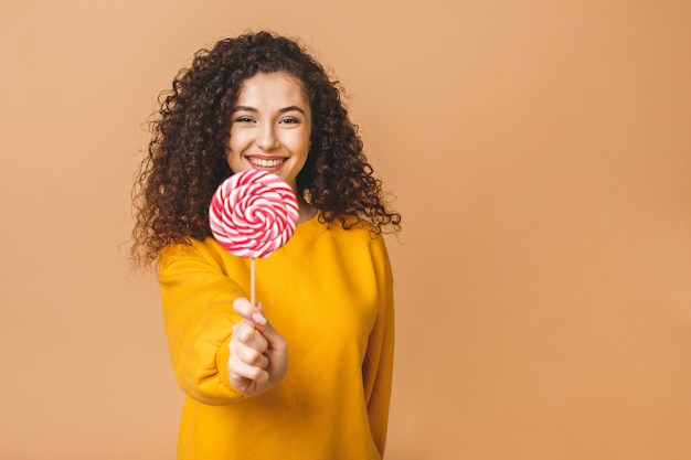 Menina encaracolada surpresa comendo pirulito. Mulher modelo da beleza que guarda os doces coloridos doces cor-de-rosa do pirulito, isolados no fundo bege.