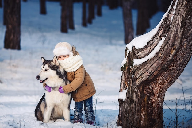 Menina engraçada abraçando seu cachorro Malamute no inverno na floresta