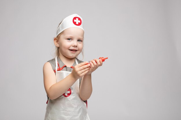 Menina engraçada bonita jogando vestindo uniforme médico segurando a seringa de brinquedo, olhando para a câmera