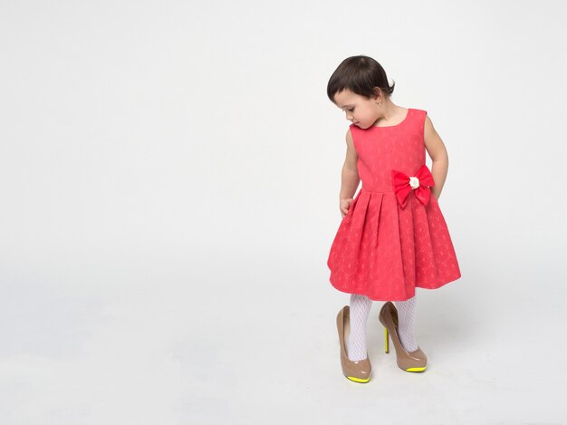 Menina engraçada com cabelo preto e um vestido bacana experimentando os sapatos de salto alto da mãe isolados