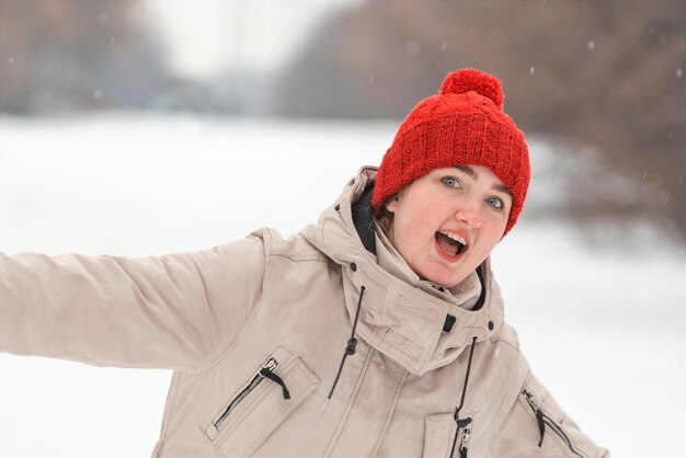 Foto menina engraçada com chapéu de tricô vermelho com bubo em fundo de parque de neve menina ri e gira lá fora mulher de roupas de inverno lá fora