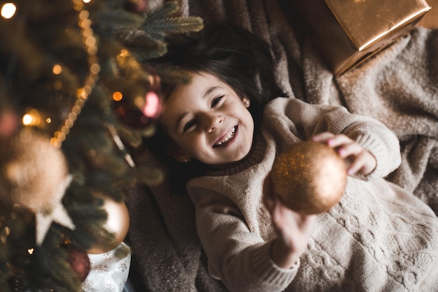 Menina engraçada e alegre segurando uma bola de Natal deitada no chão