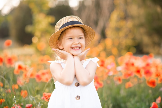 Menina engraçada e fofa usa chapéu de palha e vestido branco de verão sobre o fundo do prado de flores
