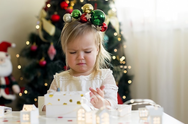 Menina engraçada em um vestido branco abrindo uma caixa de presente de Natal em casa perto da árvore de Natal