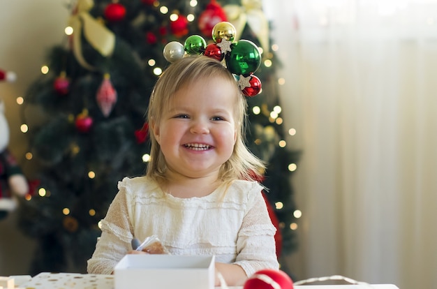 Menina engraçada em um vestido branco abrindo uma caixa de presente de Natal em casa perto da árvore de Natal