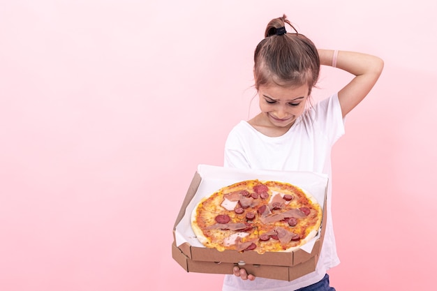 Menina engraçada olha pensativamente para pizza em uma caixa, fundo rosa, copie o espaço.