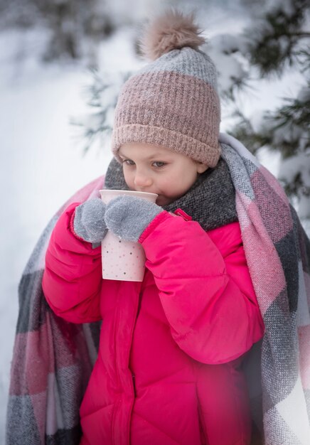 Menina enrolada em um cobertor tomando chá na floresta de inverno