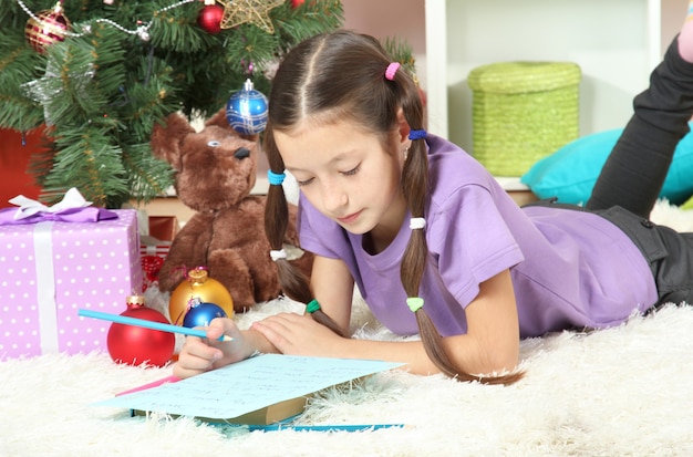 Menina escrevendo carta para o Papai Noel perto da árvore de Natal