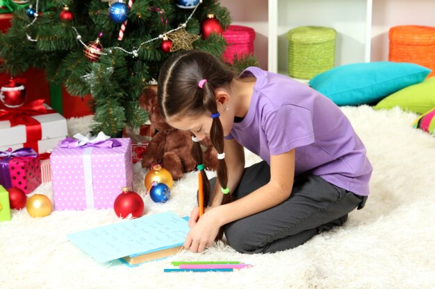 Menina escrevendo carta para o papai noel perto da árvore de natal