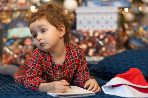 Foto menina, escrevendo uma carta para o papai noel. criança sonha perto da árvore de natal sobre desejo, presente no ano novo