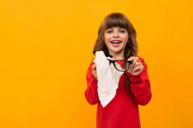 Menina esfrega óculos com um guardanapo em uma parede laranja