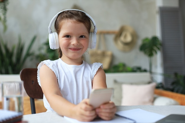 Menina esperta em fones de ouvido fazer lição de casa online aula no smartphone, criança pequena em fones de ouvido estudar na Internet, ter webconferência ou aula durante a quarentena.