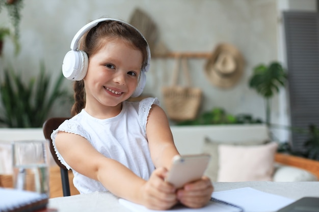 Menina esperta em fones de ouvido fazer lição de casa online aula no smartphone, criança pequena em fones de ouvido estudar na Internet, ter webconferência ou aula durante a quarentena.