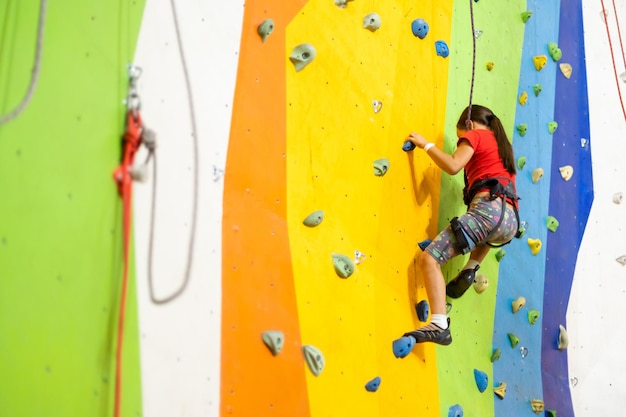 Menina esportiva escalando pedra artificial em uma parede prática no ginásio