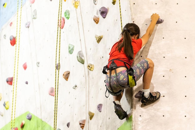 Menina esportiva escalando pedra artificial em uma parede prática no ginásio