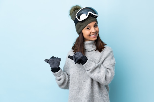 Menina esquiadora de raça mista com óculos de snowboard sobre azul isolado apontando para o lado para apresentar um produto.