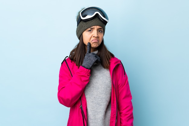 Menina esquiadora de raça mista com óculos de snowboard sobre parede azul isolada, tendo dúvidas enquanto olha para cima.