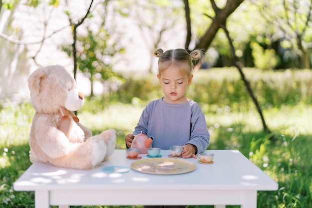 Menina está em uma mesa no jardim e joga festa de chá com um ursinho de pelúcia