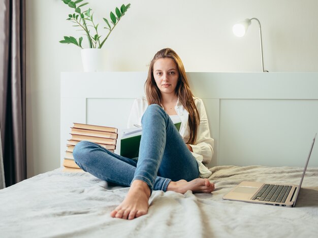 Menina está estudando enquanto está sentado em uma sala iluminada em casa no sofá ao lado de uma pilha de livros.