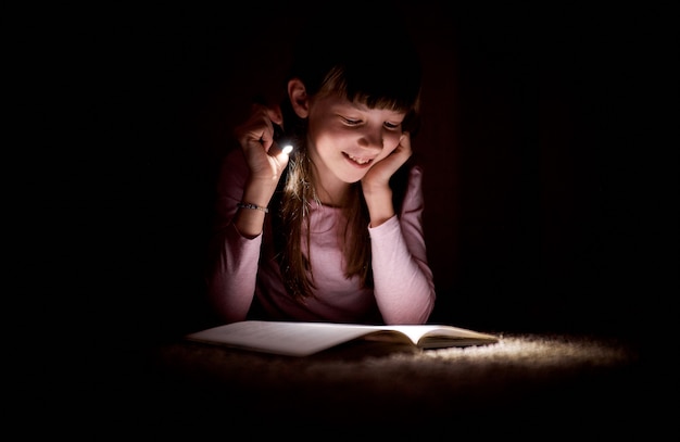 Menina está lendo um livro com uma lanterna em um quarto escuro à noite.