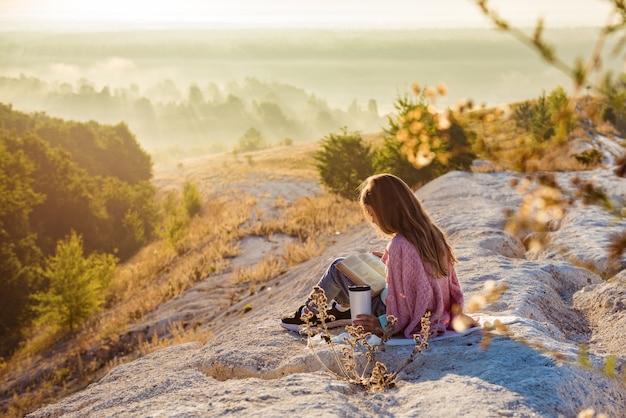 Menina está lendo um livro, enquanto está sentado contra um belo cenário da natureza. Ela está segurando um livro nas montanhas. Férias de verão e conceito de estilo de vida