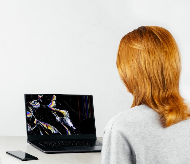 menina está sentada a uma mesa e olhando para um laptop com uma tela quebrada e rachada em pontos coloridos