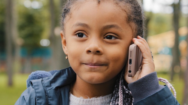 Menina étnica falando no telefone celular ao ar livre retrato engraçado criança multirracial atender chamada telefônica pequeno bebê afro-americano filha estudante em pé na rua conversando com smartphone