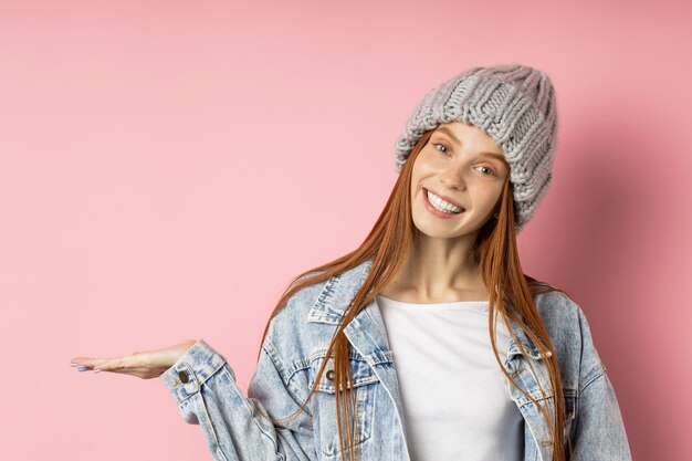 Menina europeia satisfeita com cabelo ruivo em uma jaqueta jeans grande, chapéu cinza de inverno, mantendo a palma da mão levantada, segurando o espaço da cópia, anunciando seu produto isolado de fundo rosa. Pessoas, conceito de promoção.