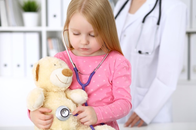 Menina examinando seu ursinho de pelúcia pelo estetoscópio conceito de confiança do paciente infantil de cuidados de saúde