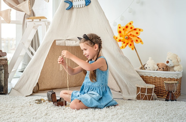 Menina explorando joias em um pequeno baú sentada no chão do quarto em frente à cabana