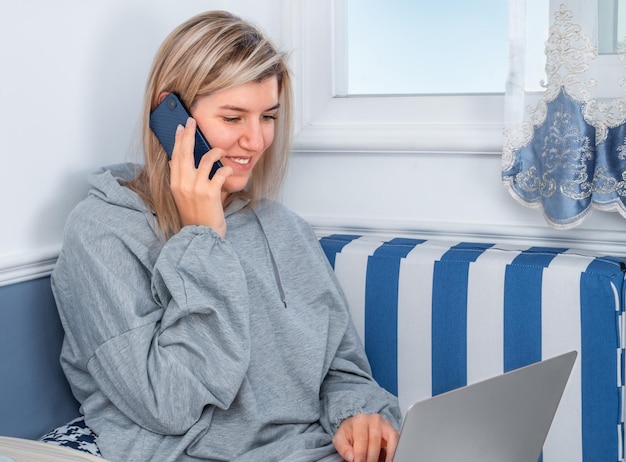 Menina falando no telefone com um sorriso e usando um laptop perto da janela em casa