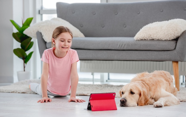 Menina fazendo alongamento com aulas de ioga online no tablet e ficando no chão com o cachorro golden retriever