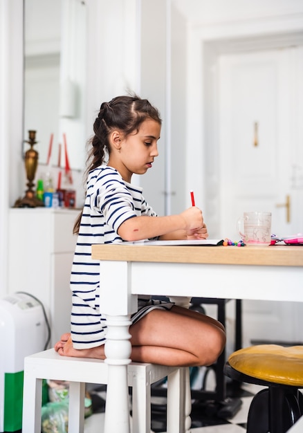 Foto menina fazendo desenhos em um livro enquanto está sentada à mesa em casa