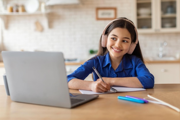 Menina fazendo lição de casa no laptop usando fones de ouvido tomando notas dentro de casa