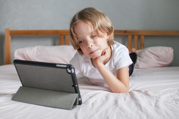 Menina fazendo lição de casa online, deitada na cama em casa. Comunicação com parentes online. O conceito de início de um novo ano letivo. Foto de alta qualidade